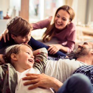 A family of four—a father, a mother, and two children—are gathered by a couch that the father is laying on.