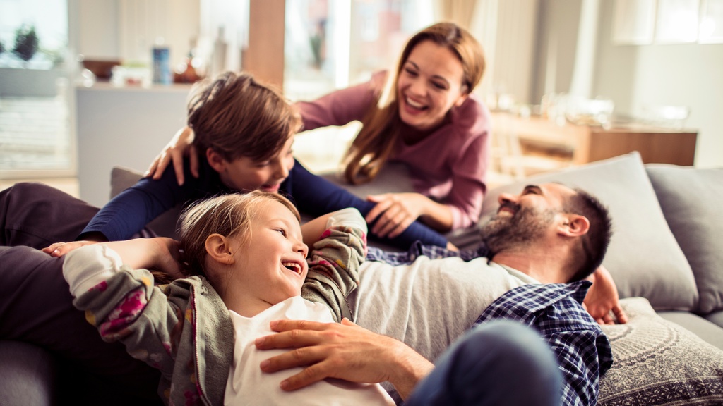 A family of four—a father, a mother, and two children—are gathered by a couch that the father is laying on.