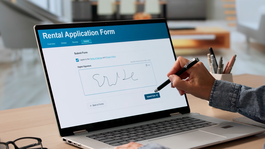 A close-up of a hand holding a stylus pen to digitally sign inside a black square on a laptop monitor on a desk.