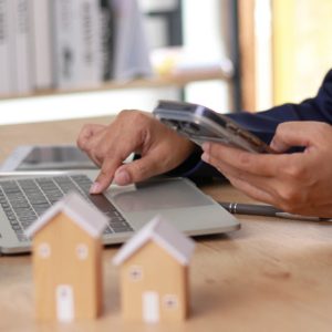 A man using a laptop while holding a smartphone in the other hand, with two miniature home models next to him.