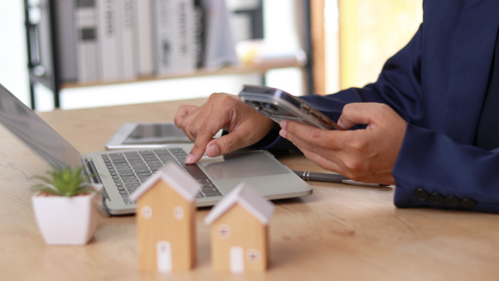 A man using a laptop while holding a smartphone in the other hand, with two miniature home models next to him.
