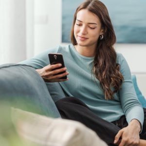 A woman is sitting on a couch while she is holding her phone. There is a plant in the background an done in the foreground.