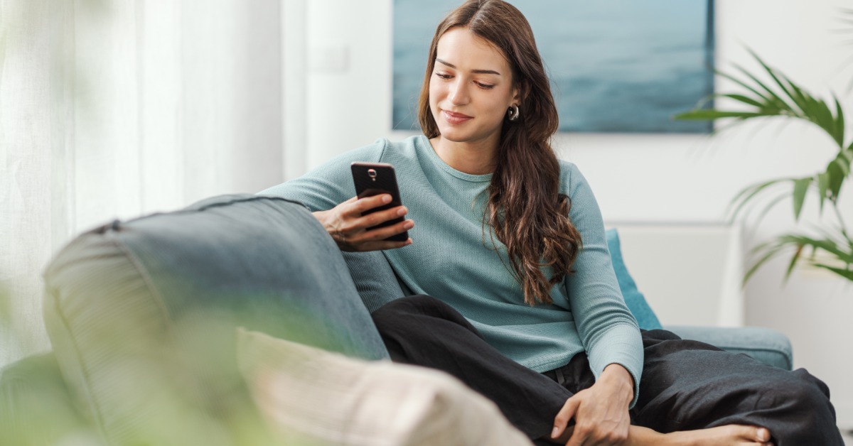 A woman is sitting on a couch while she is holding her phone. There is a plant in the background an done in the foreground.