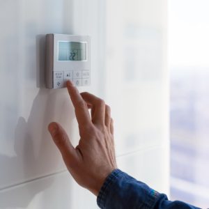 A white wall-mounted thermostat is attached to a white wall. A person is using their index finger to press a button.