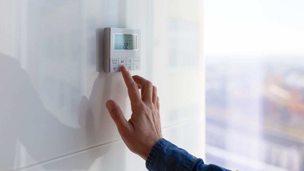 A white wall-mounted thermostat is attached to a white wall. A person is using their index finger to press a button.