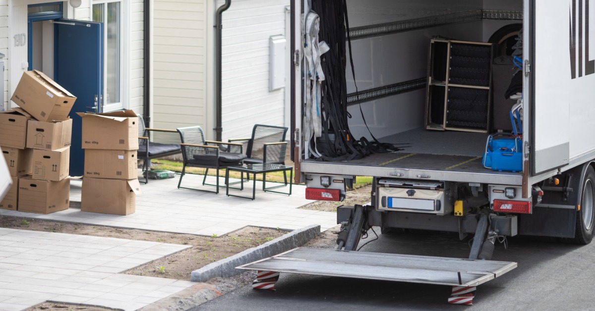 A box truck has it’s back door open. There are several items inside the truck and boxes are stacked next to it.