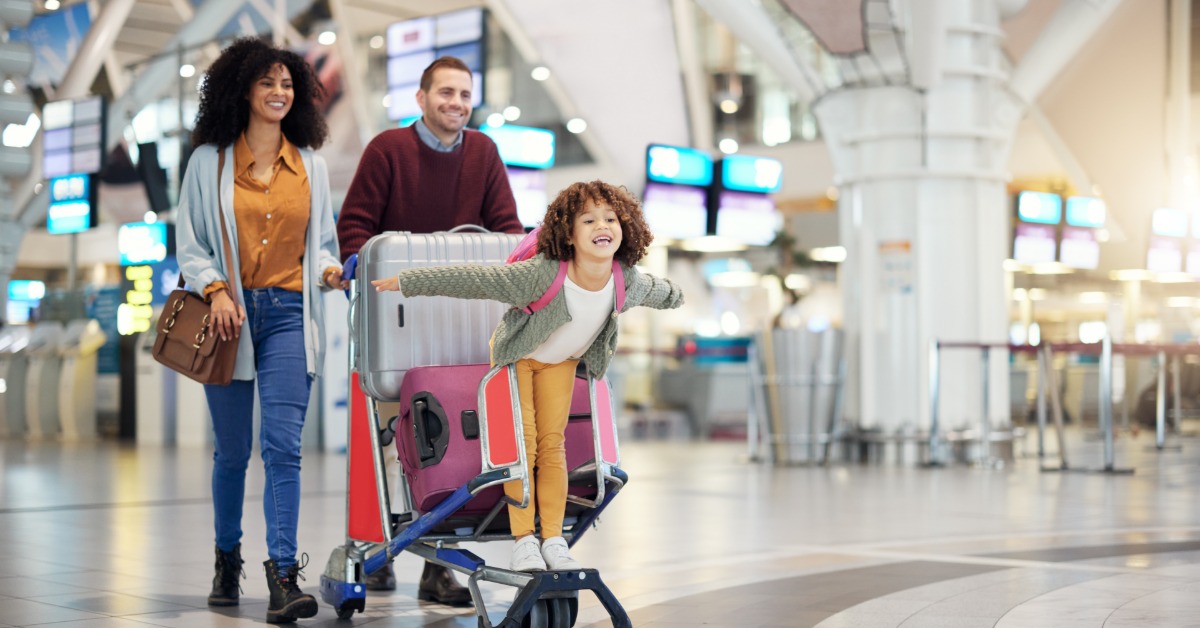 A father is pushing a cart filled with luggage through an airport. A young girl is on the front with her arms out.