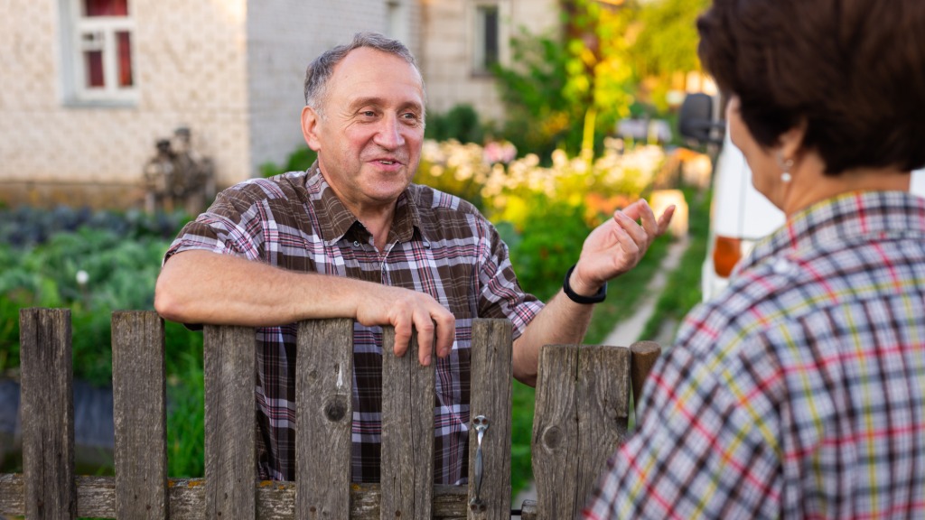 A smiling man talks with a woman over a wooden gate beside a green garden and a stone house on a sunny afternoon.