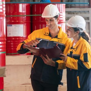 Two workers in white hard hats stand in front of large red drums. A large label is blurred on the side of the drums.