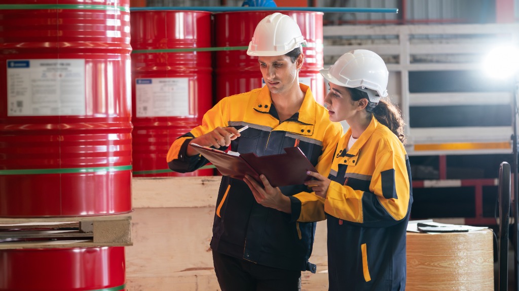 Two workers in white hard hats stand in front of large red drums. A large label is blurred on the side of the drums.