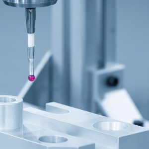 A CMM probe with a ruby tip and a short stem hovers over an aluminum part with a smooth surface and several holes.