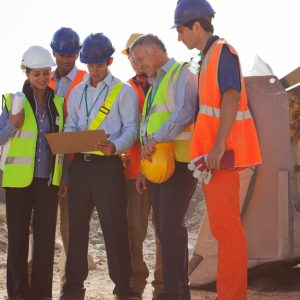 Seven construction crew members standing on a jobsite. One crew member holds a clipboard everyone looks at.
