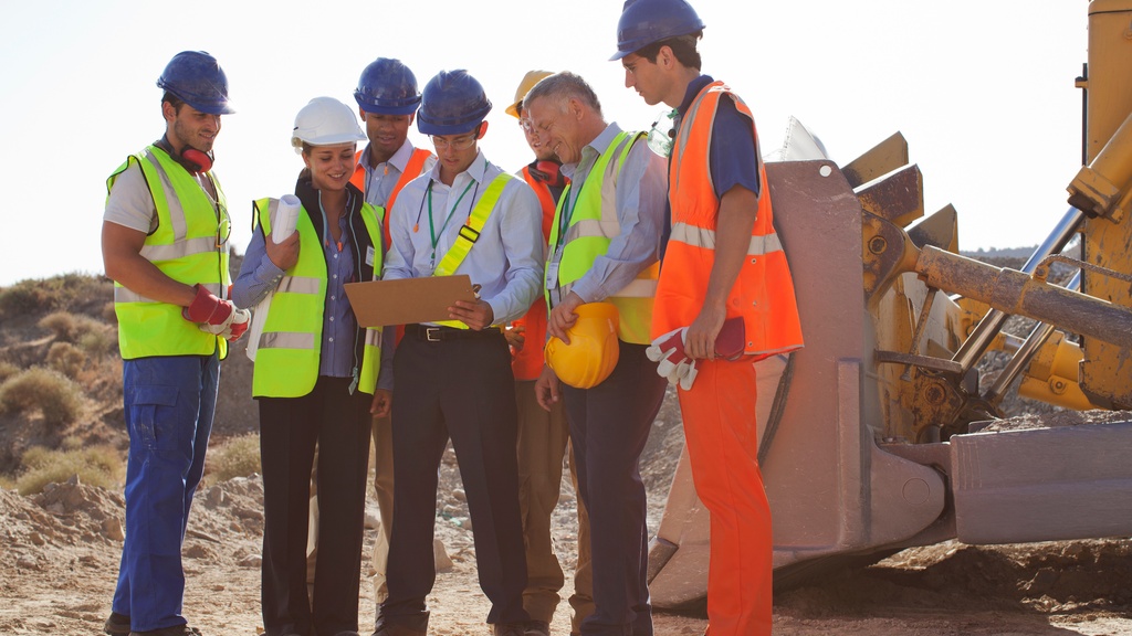 Seven construction crew members standing on a jobsite. One crew member holds a clipboard everyone looks at.