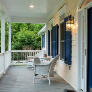 A blue front door and a glowing lantern frame a white porch with wicker chairs, shutters, flowers, and greenery.