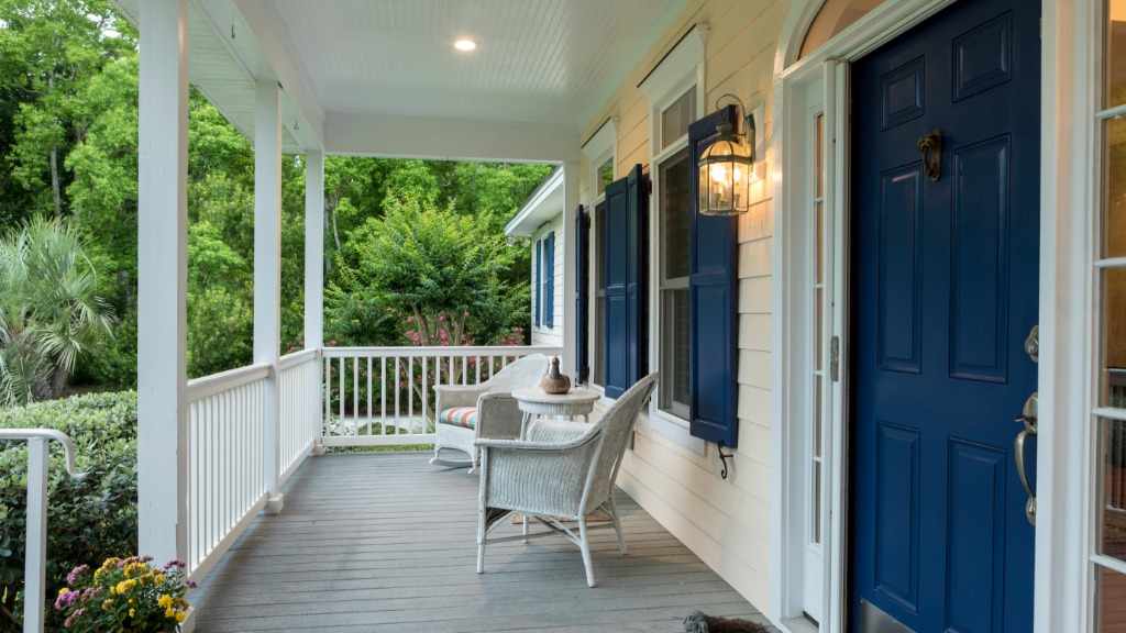 A blue front door and a glowing lantern frame a white porch with wicker chairs, shutters, flowers, and greenery.