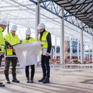 A group of workers wearing hard hats and safety vests review blueprints together at an outdoor construction site.