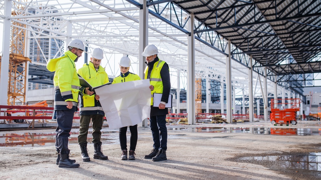 A group of workers wearing hard hats and safety vests review blueprints together at an outdoor construction site.