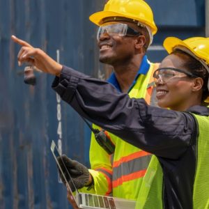 Two workers wearing yellow hard hats and holding a laptop pointing beside blue shipping containers.