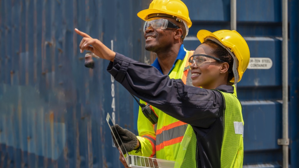 Two workers wearing yellow hard hats and holding a laptop pointing beside blue shipping containers.