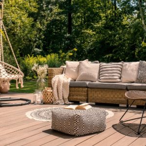 A cozy outdoor deck with wicker seating, patterned cushions, a hanging chair, potted plants, and layered decor surrounded by lush greenery.