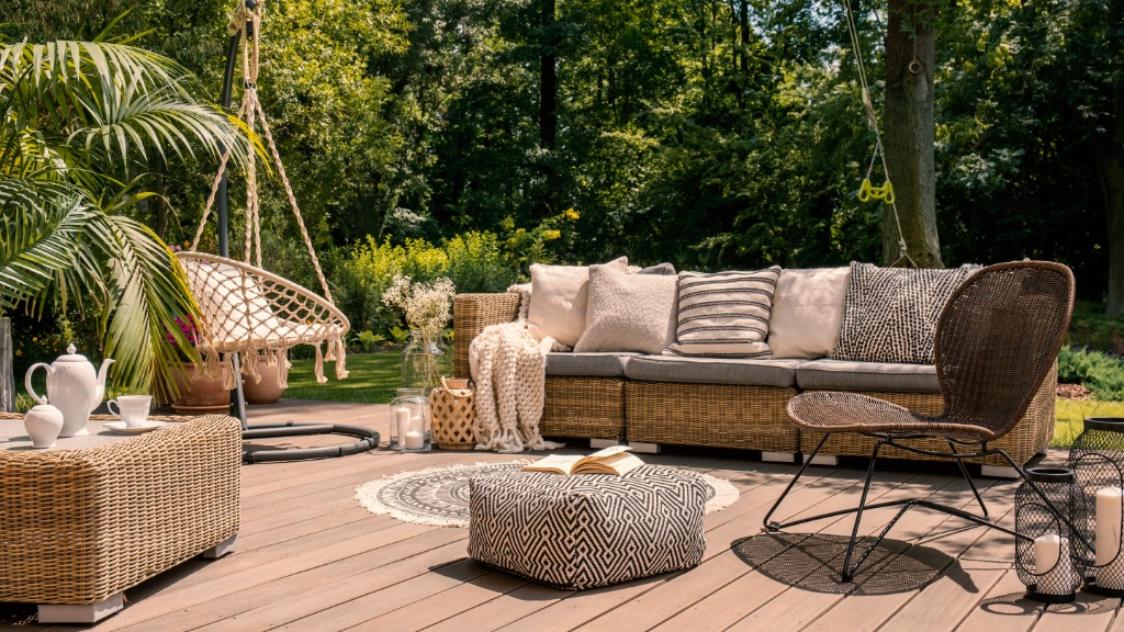 A cozy outdoor deck with wicker seating, patterned cushions, a hanging chair, potted plants, and layered decor surrounded by lush greenery.