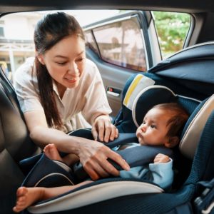 Infant rests in a rear car seat while a woman reaches inside to adjust the child's padded safety harness.