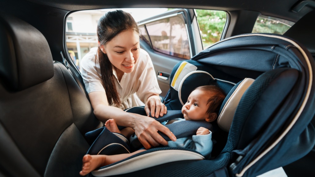 Infant rests in a rear car seat while a woman reaches inside to adjust the child's padded safety harness.