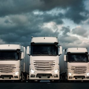 Five massive white trucks parked beside each other underneath a dark, cloudy sky. All truck headlights are activated.