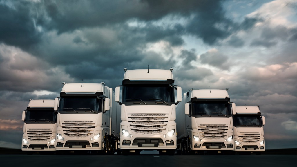 Five massive white trucks parked beside each other underneath a dark, cloudy sky. All truck headlights are activated.