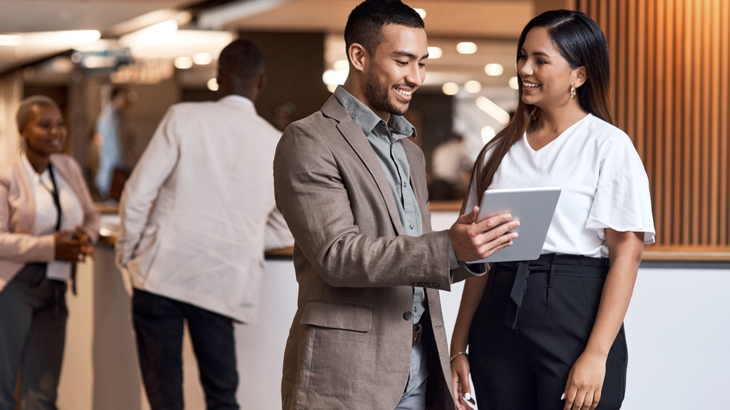 A professionally dressed person at a trade show holds a tablet in front of a guest. The duo stand by two more guests.