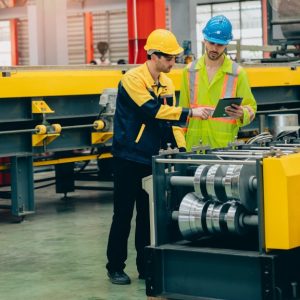 Two engineers wearing hard hats look at a small tablet in a large, brightly lit manufacturing facility.