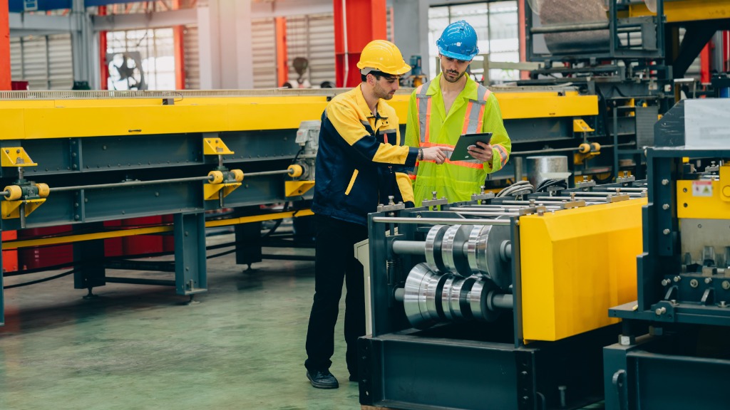 Two engineers wearing hard hats look at a small tablet in a large, brightly lit manufacturing facility.