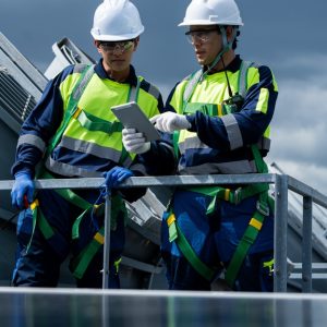 Two technicians wearing white hard hats looking at a tablet outside an industrial facility. Gray clouds are in the sky.