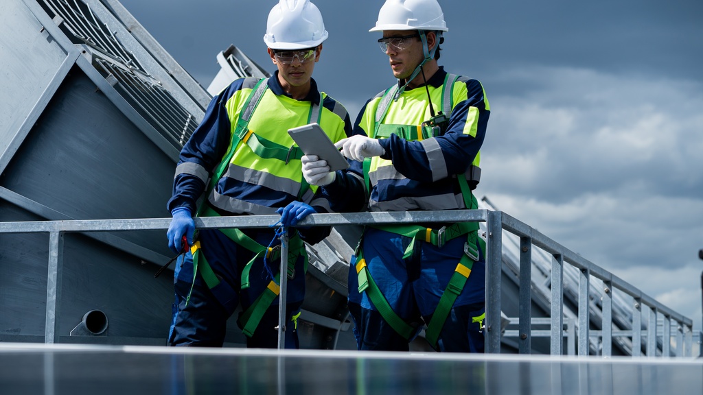 Two technicians wearing white hard hats looking at a tablet outside an industrial facility. Gray clouds are in the sky.