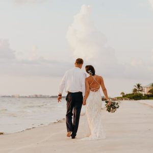 A man and woman in wedding attire walking barefoot along a sandy beach beside calm water under a cloudy sky.