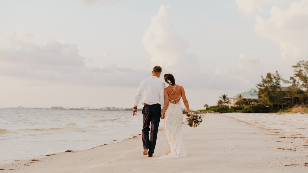A man and woman in wedding attire walking barefoot along a sandy beach beside calm water under a cloudy sky.