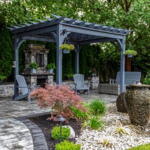 Landscaped backyard with a gray pergola, stone patio, seating area, and decorative water urns surrounded by greenery.