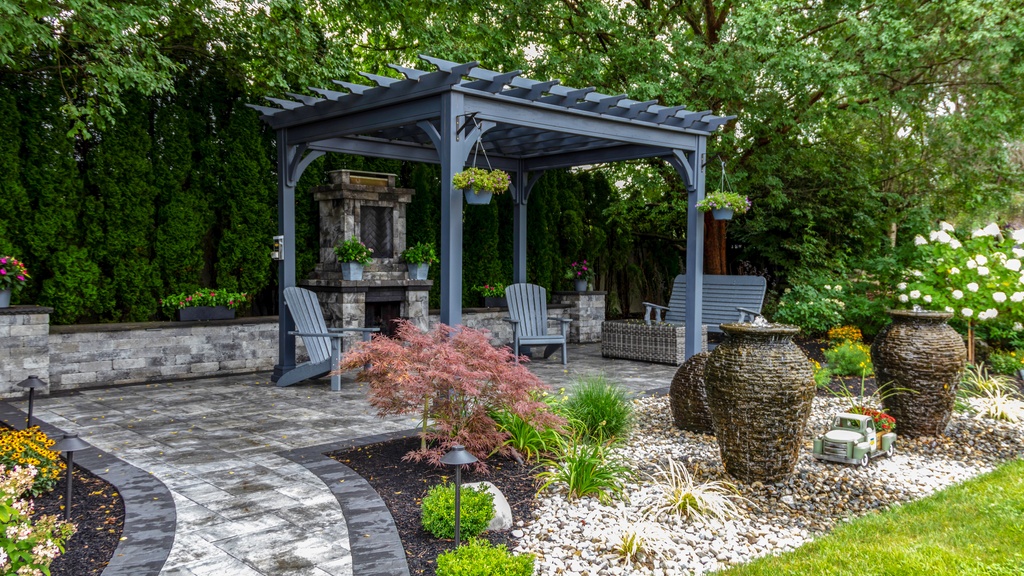 Landscaped backyard with a gray pergola, stone patio, seating area, and decorative water urns surrounded by greenery.