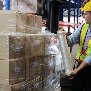 A man in a high-visibility vest and a hard hat wrapping a pallet of cardboard boxes with stretch film.