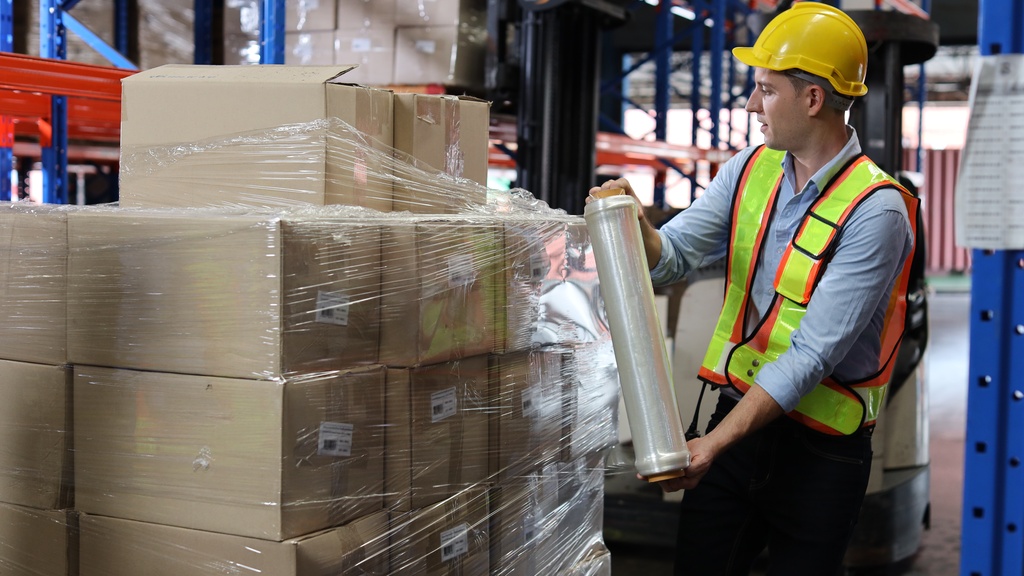 A man in a high-visibility vest and a hard hat wrapping a pallet of cardboard boxes with stretch film.