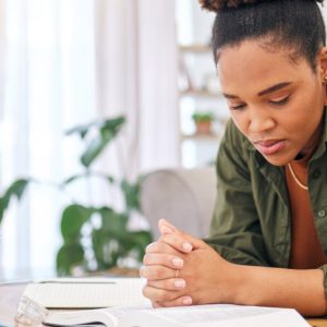 A woman sits at a table and reads the Bible with her hands clasped. A notebook, glasses, and a potted plant are nearby.