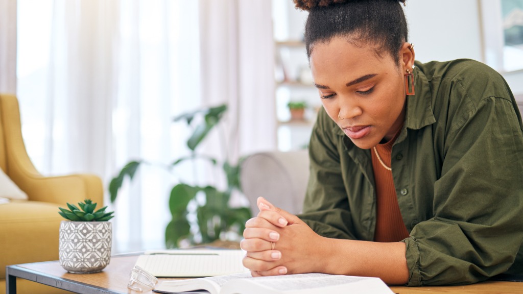 A woman sits at a table and reads the Bible with her hands clasped. A notebook, glasses, and a potted plant are nearby.