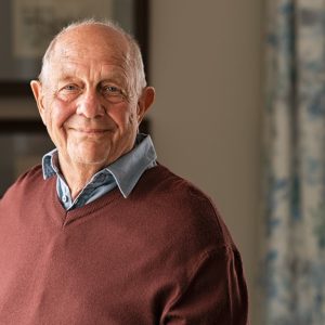 A senior man wearing a brown sweater while smiling and sitting inside a room with floral curtains and artwork.
