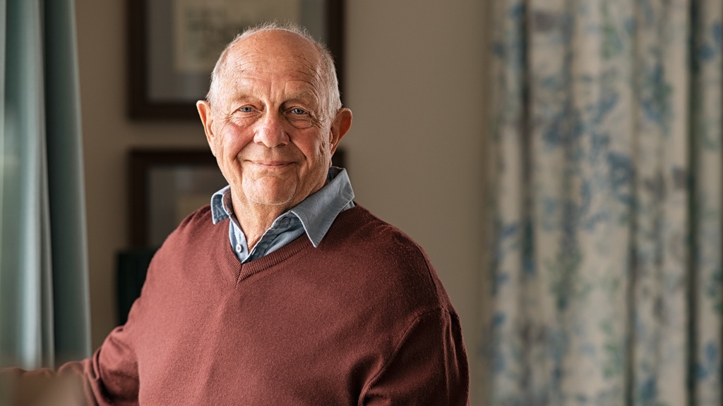 A senior man wearing a brown sweater while smiling and sitting inside a room with floral curtains and artwork.