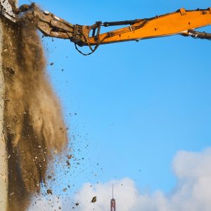An excavator digging into the side of a building during the daytime. Debris falls from where the machine makes contact.