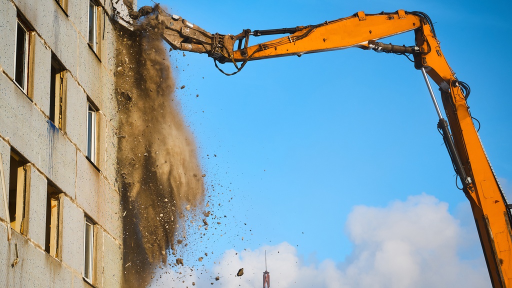 An excavator digging into the side of a building during the daytime. Debris falls from where the machine makes contact.